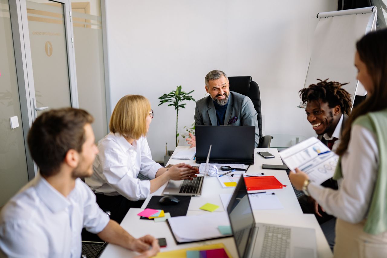 Four colleagues engaged in conversation at their office workspace, surrounded by desks and office supplies.